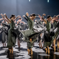 Grupa tancerek w zielonych sukniach i w podobnych tanecznych pozach, z uniesionymi rękoma. Jednolite, szare tło.