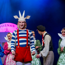 Aktor przebrany za Koziołka Matołka, w czerwonych spodniach na szelkach i biało-niebieskiej bluzce, za nim kilka osób w strojach ludowych.