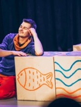 Uśmiechnięty mężczyzna klęcząc podpiera się o drewniane pudło z rusunkiem ryby, obok stoi kolejne pudło z rysunkiem fal.