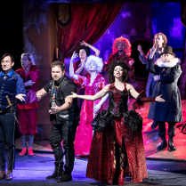 Zdjęcie ze spektaklu - na scenie aktorzy w różnorodnych strojach; na pierwszym planie aktorka w długiej balowej sukni, obok niej mężczyzna w skórzanych spodniach, skórzanej kamizelce i T-shircie.