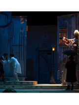 Na zdjeciu czworo aktorów w dziewiętnastowiecznych strojach na scenie. Po lewej stronie dwaj mężczyźni stoją pochyleni ku sobie, po prawej na balkonie widoczna kobieta w długiej sukni, przed nią na chodniku stoi mężczyzna w czarnym płaszczu i kapeluszu pod parasolem.