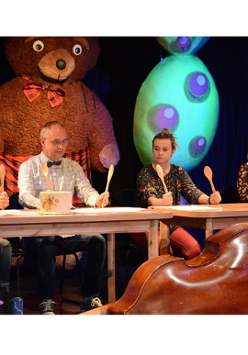 Na scenie czworo aktorów siedzi za stołem. W rękach trzymają uniesione drewniane łyżki, a na stołach leżą odwrócone do góry dnem garnki.