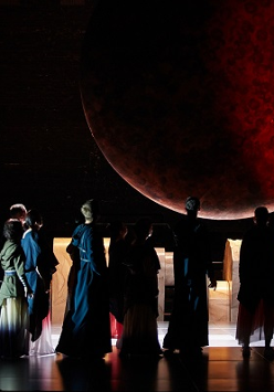 Ciemne zdjęcie sceny, nad którą unosi się duże czerwono-czarne koło przypominające księżyc. Na scenie aktorzy odwróceni tyłem do widzów i zwróceni w kierunku czerwonego koła. W tle elementy scenografii.