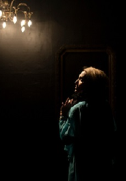 Ciemne zdjęcie, na którym widać kobietę odwróconą bokiem, z oświetloną twarzą. U góry zdjęcia świecąca się lampa w formie ozdobnego żyrandola.