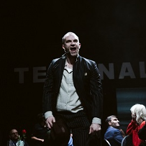 Mężczyzna, który stoi z otwartymi ustami, wokół niego siedzące na krzesłach osoby. Ciemne tło.