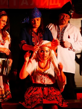 Aktorzy w strojach ludowych: jedna kobieta siedzi, druga nakłada jej białą chustę na głowę. Pozostałe osoby obserwują to.