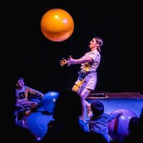 Kobieta na scenie podrzuca dużą pomarańczową piłkę. Widocznych jest kilka obserwujących ją osób. Ciemne tło.