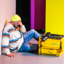 na zdjęciu mężczyzna siedzi pod ścianą, ręką zasłania twarz, przed nim na podłodze leży żółta torba termiczna do przewozu jedzenia