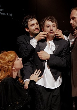 Dwóch mężczyzn soi blisko trzeciego mężczyzny, jeden z nich dotyka jego twarzy. Dwie kobiety przygladają się mężczyznom.