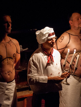 Na zdjęciu trzech mężczyzn: jeden, w środku, ubrany w biały uniform z biała kucharską czapką na głowie, trzyma w ręce świecznik, dwóch pozostałych z nagimi wytatułowanymi torsami za nim.