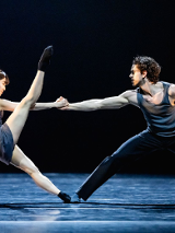 Na scenie kobiet ai mężczyzna w trakcie tańca baletowego. Kobieta stoi naprzeciwko mężczyzny, trzyma go za rękę, jedną nogę podnosi wysoko do góry. Mężczyna trzyma ją mocno, pochylając się do przodu..