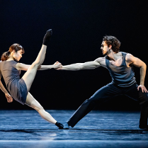 Na scenie kobiet ai mężczyzna w trakcie tańca baletowego. Kobieta stoi naprzeciwko mężczyzny, trzyma go za rękę, jedną nogę podnosi wysoko do góry. Mężczyna trzyma ją mocno, pochylając się do przodu..