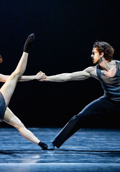 Na scenie kobiet ai mężczyzna w trakcie tańca baletowego. Kobieta stoi naprzeciwko mężczyzny, trzyma go za rękę, jedną nogę podnosi wysoko do góry. Mężczyna trzyma ją mocno, pochylając się do przodu..