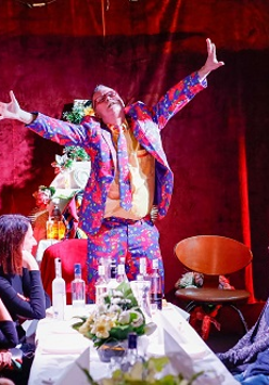 Przy stole siedzi kilka osób, u szczytu stołu stoi mężczyzna w kolorowym garniturze z szeroko rozłożonymi rękoma. Na stole kilka butelek alkoholu.