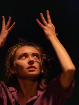 Kobieta w teatralnej pozie z uniesionymi rękoma do góry, ciemne tło.