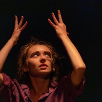 Kobieta w teatralnej pozie z uniesionymi rękoma do góry, ciemne tło.