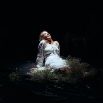 Na czarnym tle zdjęcie młodej, uśmiechniętej dziewczyny w białej sukni. Dziewczyna siedzi oparta rękoma o podłogę, patrzy ku górze.