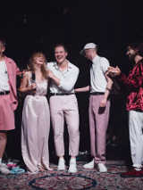 Siedmioro ludzi ubrani w odcienie różowego stoją na scenie wyłozonej dywanami. Za nimi instrumenty muzyczne.