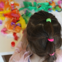 Zdjęcie zrobione z góry. Dziewczynka stoi tyłem do obiektywu. Przed nią na stoliku leżą kolorowe przybory plastyczne.