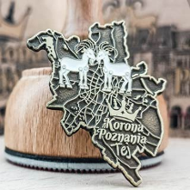 Metalowa przypinka w kształcie mapy Poznania z wizerunkiem dwóch koziołków i napisem "Korona Poznania Tej", oparta o drewniane naczynie. Tło rozmyte.