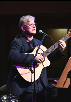 Zdjęcie mężczyzny na scenie, na której gra na gitarze i śpiewa. Przed nim stoi mikrofon na statywie.