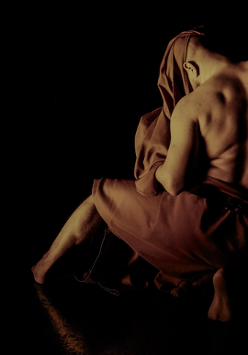 Zdjęcie przedstawiające mężczyznę z odsłoniętymi plecami i ramionami, częściowo okrytego tkaniną. Postać jest ukazana w półmroku na czarnym tle, z widocznym światłem podkreślającym mięśnie mężczyzny i fakturę materiału.