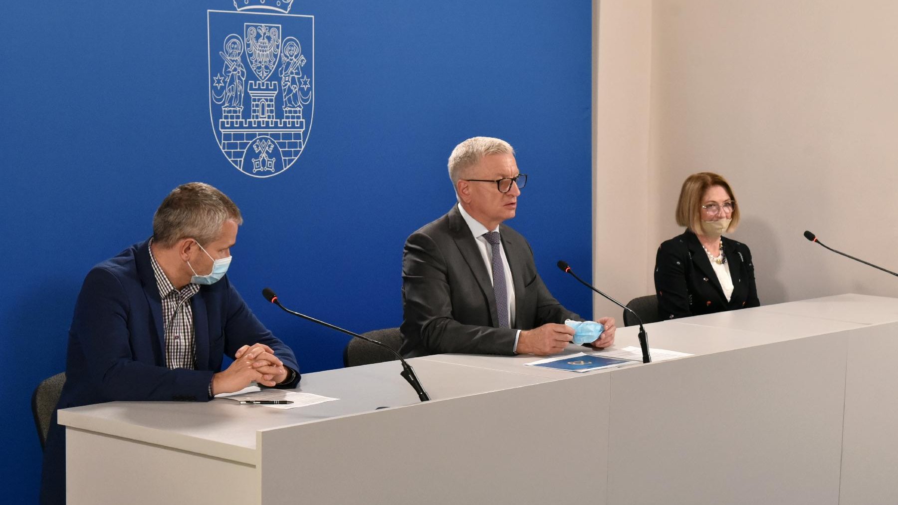 Centrum prasowe UMP: za szarym stołem, na tle ścianki z herbem miasta siedzą trzy osoby. Od lewej: Jędrzej Solarski, Jacek Jaśkowiak - mówiący do mikrofonu oraz prof. Iwona Mozer-Lisewska - grafika artykułu