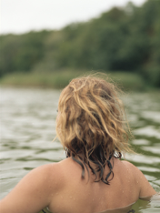 Kobieta w wodzie, widać plecy i długie włosy. Z przodu las.