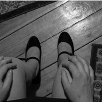 Na czarno-białym zdjęciu będącym kadrem z wystawy widzimy nogi siedzącej w spudnczce kobiety. W tle drewniana podłoga i fragmenty dwóch dywaników.