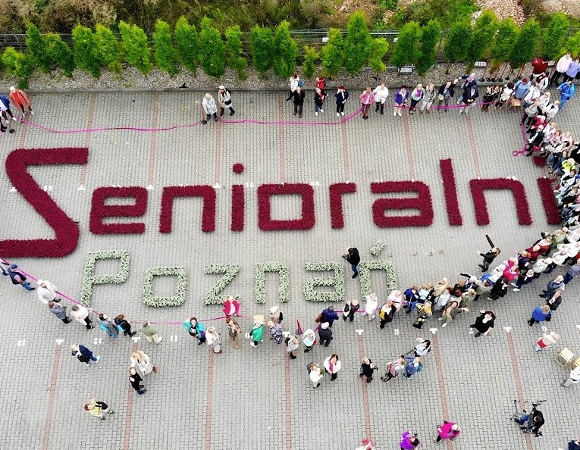 grafika przedstawia widok z góry na plac na którym ułożony został z kwiatów napis Senioralni