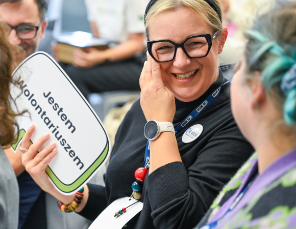 Na zdjęciu dwie uśmiechnięte kobiety, jedna trzyma tabliczkę z napisem: jestem wolontariuszem