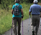 Na zdjęciu dwoje seniorów z kijkami do nordic walkingu, tyłem do obiektywu