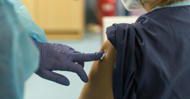 Na zdjęciu dłoń w medycznej rękawiczce przykłada wacik do miejsca po szczepieniu innej osoby - widać tylko kawałek jej pleców i ramię