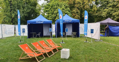Zdjęcie mobilnego punktu szczepień: trzy niebieskie namioty na trawniku, przed nimi banery informacyjne i pomarańczowe leżaki