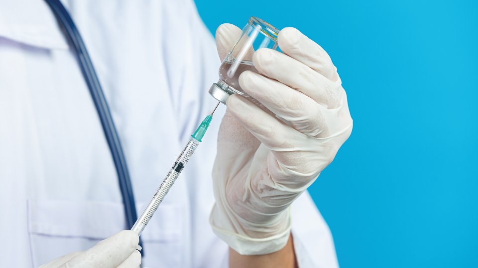 na zdjęciu w rękach medyka zabezpieczonych rękawiczkami trzymane są - fiolka z preparatem szczepiącym i strzykawka. - grafika artykułu