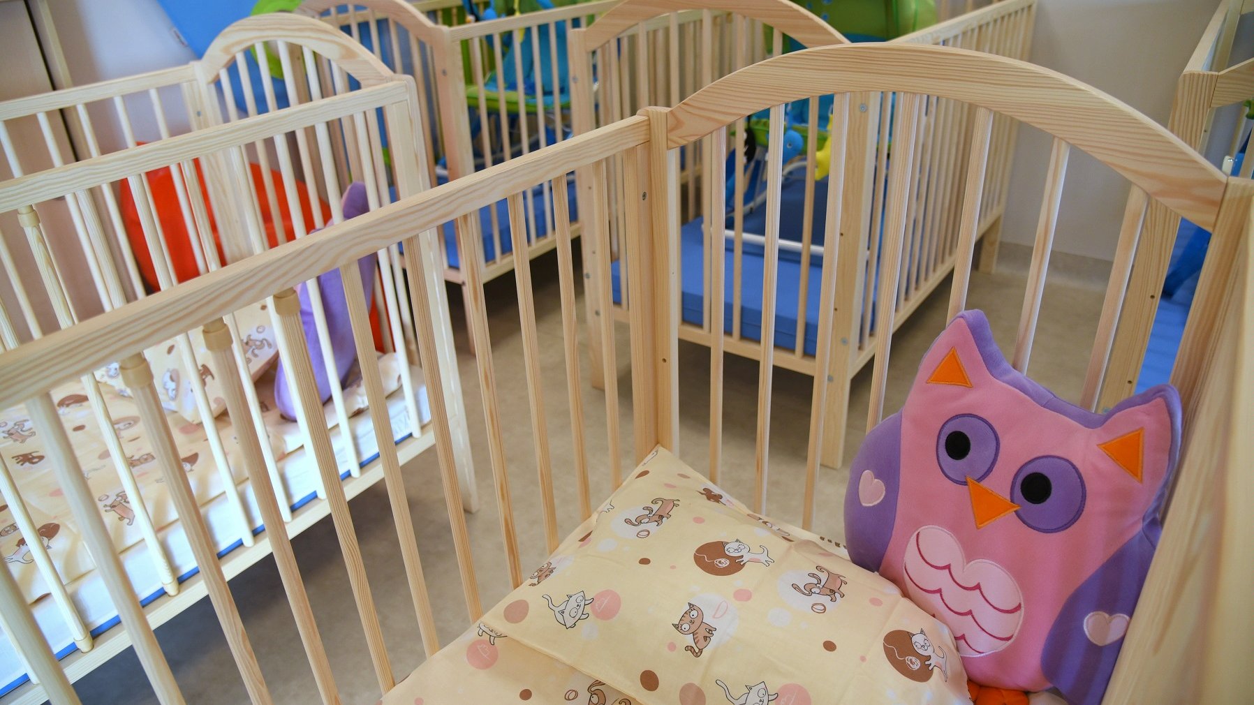 Dzieciece łóżeczka, stoją obok siebie. W nich kolorowe materace i fioletowa poduszka w kształcie sowy - grafika artykułu
