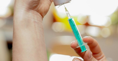 Na zdjęciu widać dwie dłonie osoby, która przygotowuje szczepionkę, w centrum zdjęcia strzykawka