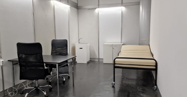 Na zdjęciu wnętrze punktu szczepień, widać stół z dwoma krzesłami i lekarską leżankę