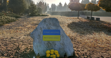 Na zdjęciu biały kamień na cmentarzu, na nim tabliczka, przed nim żółte znicze