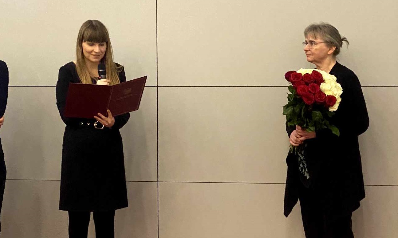 zdjęcie przedstawia Rzeczniczkę Praw Dziecka odczytującą laudację oraz pania Marzenę Affeld stojąca z kwiatami - grafika artykułu