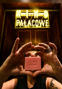 Kobieta trzyma w rękach małe różowe mydło z napisem: Kino Pałacowe. Nie widać jej twarzy. Nad nią neon kinowy.