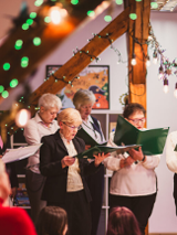 Grupa śpiewającycj osób w udekorowanym świątecznie pomieszczeniu.