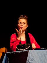 Dwie kobiety, jedna siedzi przy stole i gestykuluje, druga stojąc z założonymi rękoma patrzy w jej kierunku. Ciemne tło.