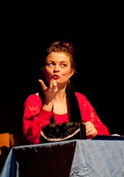 Dwie kobiety, jedna siedzi przy stole i gestykuluje, druga stojąc z założonymi rękoma patrzy w jej kierunku. Ciemne tło.