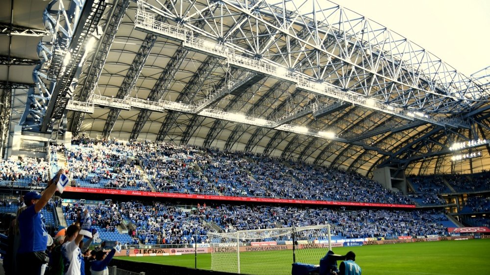 Na zdjęciu jedna z trybun Stadionu Miejskiego w Poznaniu podczas meczu Lecha Poznań. Na trybunie widać wielu wiwatujących kibiców, ubranych w niebiesko-białe barwy Lecha. Na pierwszym planie zielona murwa stadionu i jedna z bramek piłkarskich - grafika artykułu