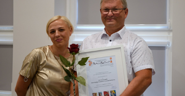 Mężczyzna i kobieta stoją obok siebie i uśmiechają się do zdjęcia, mężczyzna trzyma dyplom i różę