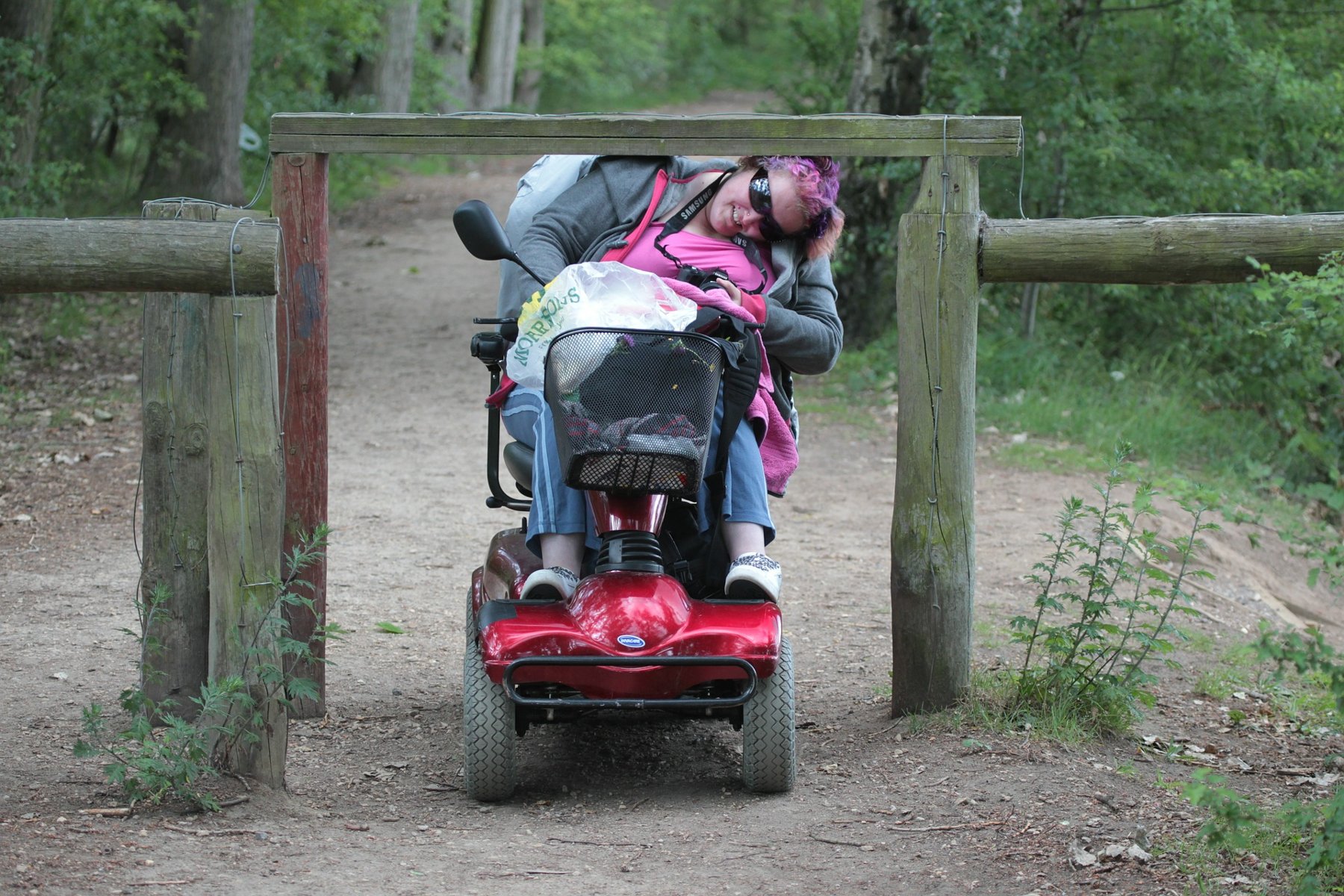 Zdjęcie przedstawia kobietę z niepełnosprawnościami na wózku elektrycznym. Przejeżdża ona pod drewnianą bramą, pochylając głowę. - grafika artykułu