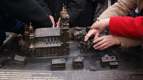 Na zdjęciu makieta Ostrowa Tumskiego w Poznaniu z Katedrą i okolicznymi budynkami, ręce dotykają drzew na makiecie, u dołu opis w alfabecie Braille'a - grafika artykułu