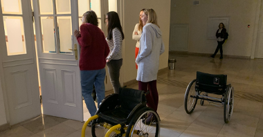 Korytarz urzędu miasta poznania. Stoi grupa osób i dwa wózki dla osón z niepełnosprawnościami, patrzą przez otwarte drzwi na schody.