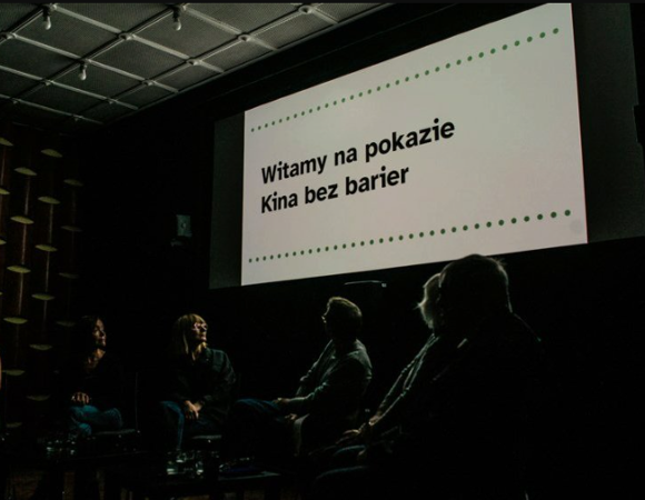 Wnętrze zaciemnionej sali kinowej. Na dużym ekranie widnieje czarny napis na białym tle: »Witamy na pokazie Kina bez barier«, ujęty w dwie poziome linie z zielonych kropek. Poniżej ekranu, w półmroku, siedzi grupa osób odwrócona tyłem lub bokiem do obiektywu, patrząca w stronę wyświetlanego komunikatu.